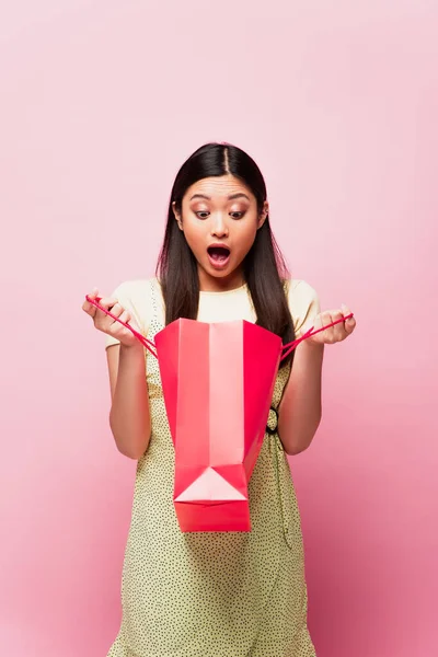 Impactado mujer asiática con la boca abierta mirando bolsa de compras en rosa - foto de stock