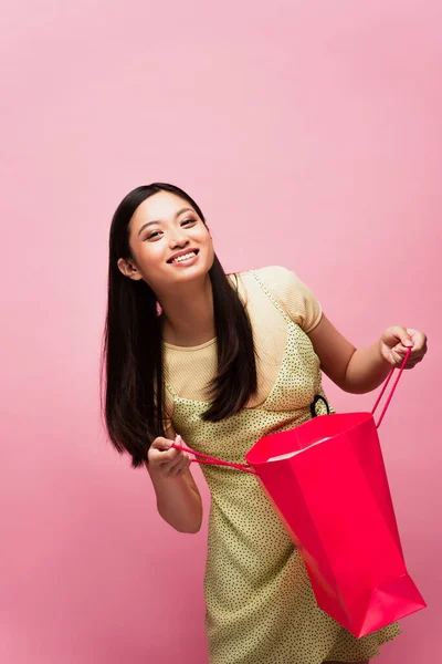 Sonriente joven asiático mujer sosteniendo bolsa aislado en rosa - foto de stock