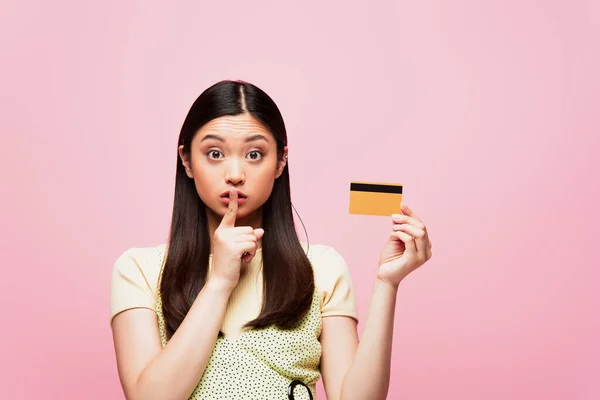 Joven asiático mujer celebración de tarjeta de crédito y mostrando silencio signo aislado en rosa - foto de stock