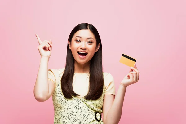 Eccitato donna asiatica punta con il dito e tenendo la carta di credito isolata su rosa — Foto stock