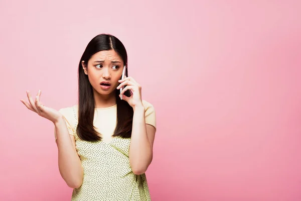 Emotional asian woman with open mouth gesturing and talking on smartphone isolated on pink — Stock Photo