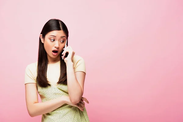 Шокована азіатська жінка з відкритим ротом говорить на смартфоні ізольовано на рожевому — стокове фото