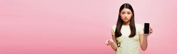 Panoramic concept of upset asian woman holding smartphone with blank screen isolated on pink — Stock Photo