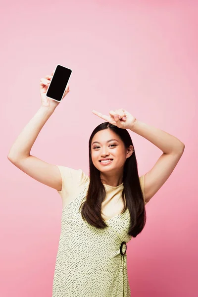Веселая азиатская девушка, указывающая пальцем на смартфон с пустым экраном, изолированным на розовый — стоковое фото