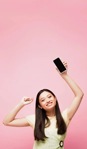 Cheerful asian girl with clenched fist holding smartphone with blank screen isolated on pink — Stock Photo