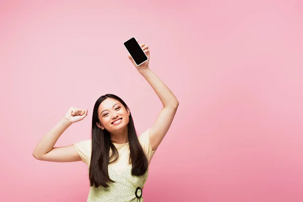 Sonriente asiático chica con apretado puño celebración smartphone con pantalla en blanco aislado en rosa - foto de stock