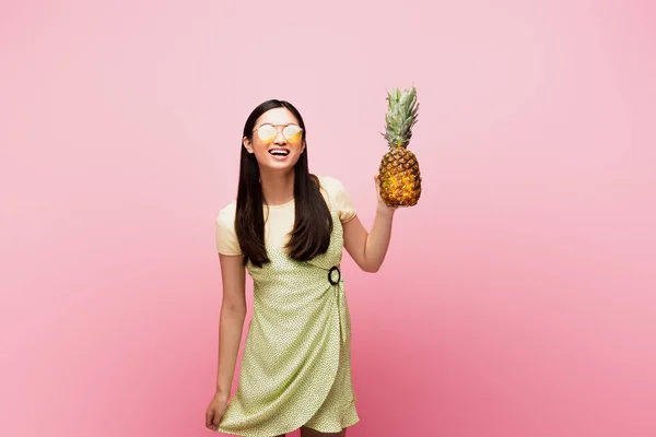 Happy asian girl in sunglasses holding ripe pineapple isolated on pink — Stock Photo