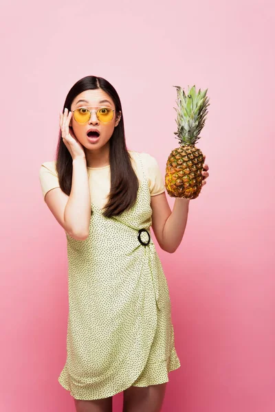 Sorprendida chica asiática en gafas de sol celebración de piña fresca en rosa - foto de stock