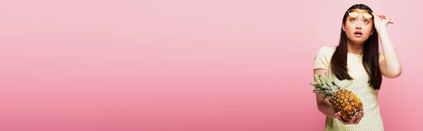 Горизонтальне зображення здивованої азіатки в сонцезахисних окулярах, дивлячись вгору і тримаючи свіжий ананас на рожевому — стокове фото