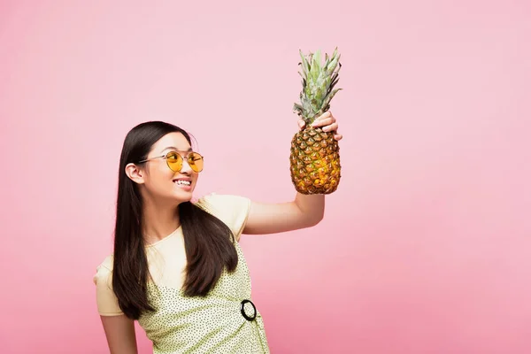 Усміхнена азіатська жінка в сонцезахисних окулярах дивиться на свіжий ананас ізольовано на рожевому — стокове фото