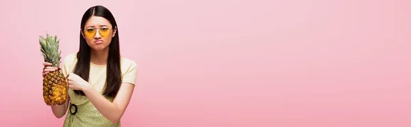 Concetto orizzontale di donna asiatica triste in occhiali da sole con ananas fresco isolato su rosa — Foto stock
