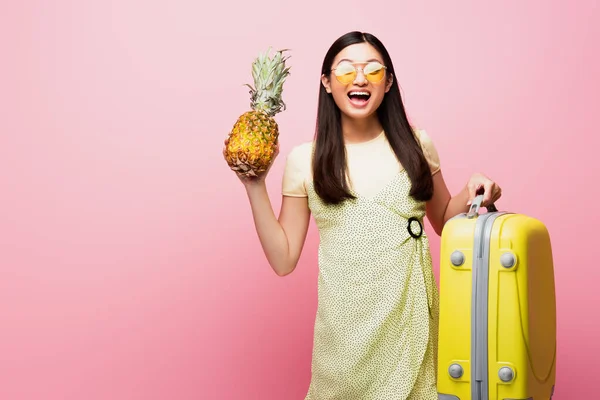 Happy asian girl in sunglasses holding baggage and fresh pineapple on pink — Stock Photo