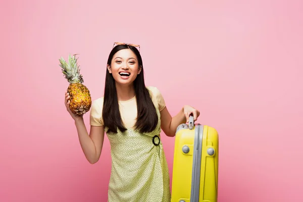 Glückliche asiatische Mädchen halten Gepäck und frische Ananas isoliert auf rosa — Stockfoto