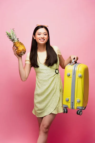 Счастливая азиатка с багажом и свежим ананасом на розовом — стоковое фото