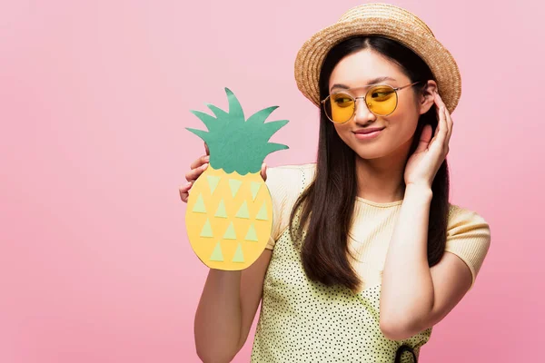 Alegre ásia menina no sol óculos e palha chapéu olhando para papel abacaxi isolado no rosa — Fotografia de Stock