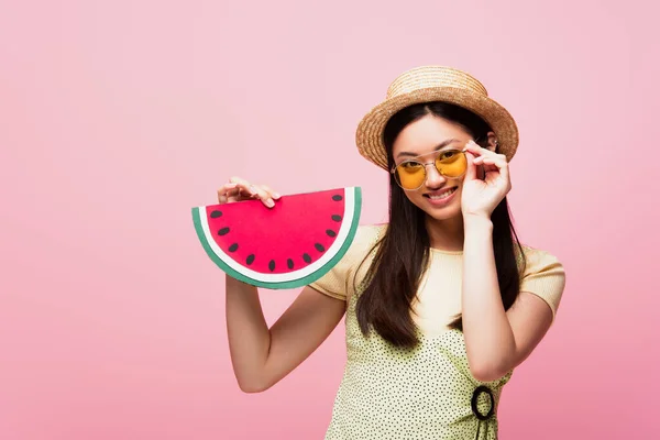 Feliz asiático chica en paja sombrero tocando gafas de sol y celebración de papel sandía aislado en rosa - foto de stock