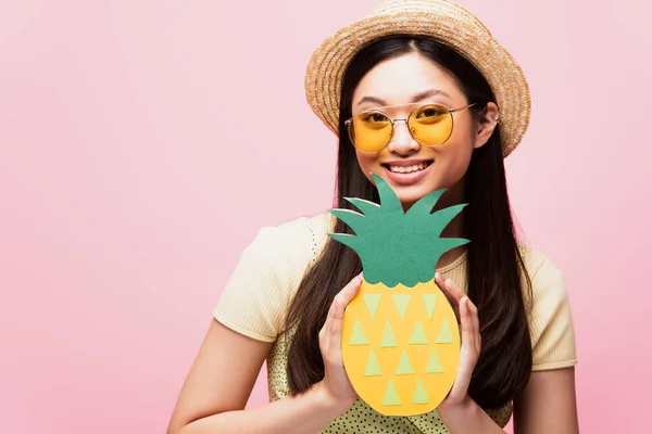 Весела азіатська жінка в сонцезахисних окулярах і солом'яному капелюсі тримає паперовий ананас ізольовано на рожевому — стокове фото
