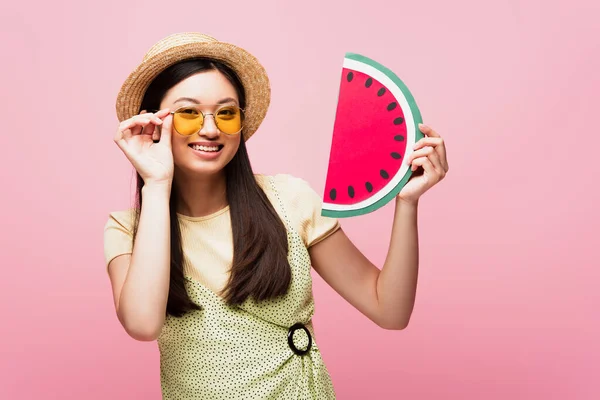 Cheerful asian girl in straw hat touching sunglasses and holding paper watermelon isolated on pink — Stock Photo