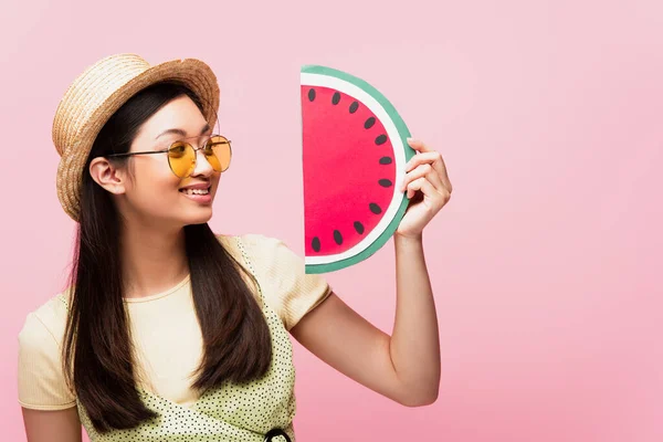 Cheerful asian girl in sunglasses and straw hat looking at paper watermelon isolated on pink — Stock Photo