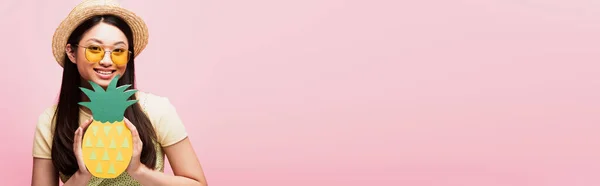 Панорамний урожай веселої азіатської дівчини в сонцезахисних окулярах і солом'яному капелюсі, що тримає паперовий ананас ізольований на рожевому — стокове фото