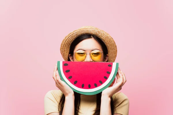 Asiático chica en gafas de sol y paja sombrero cubierta cara con papel sandía aislado en rosa — Stock Photo