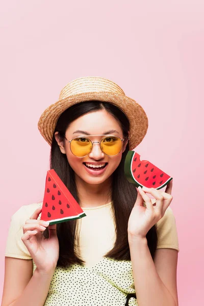 Позитивна азіатська дівчина в сонцезахисних окулярах і солом'яному капелюсі тримає нарізаний паперовий кавун ізольований на рожевому — стокове фото