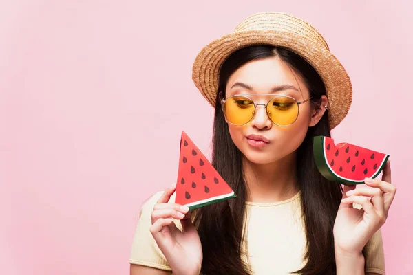 Pensive asian girl in sunglasses and straw hat holding sliced paper watermelon isolated on pink — Stock Photo