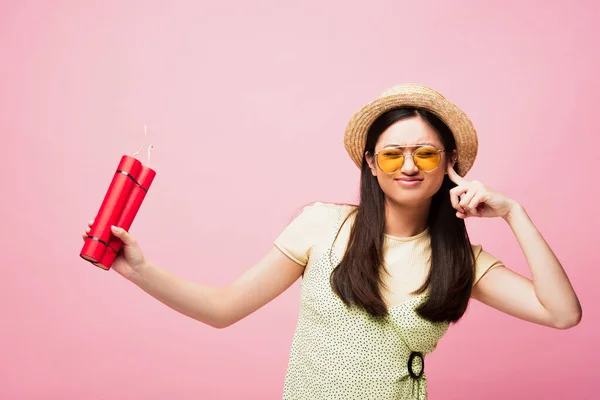 Asian girl in sunglasses and straw hat covering ear and holding dynamite isolated on pink — Stock Photo