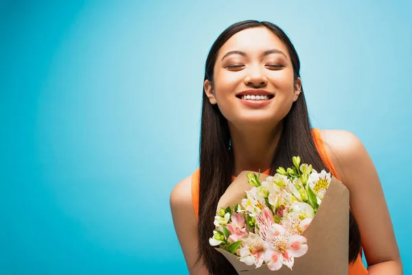 Happy asian girl with closed eyes smiling and holding flowers on blue — Stock Photo