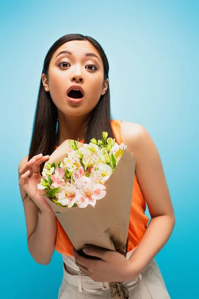Überrascht asiatisches Mädchen schaut in die Kamera und hält Blumen auf blau — Stockfoto