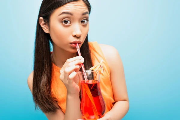 Asian girl holding glass and drinking cocktail through straw on blue — Stock Photo