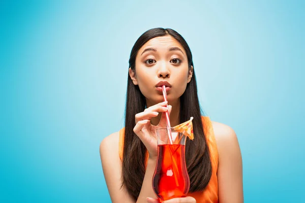 Atractivo asiático chica celebración de vidrio y beber cóctel a través de paja en azul - foto de stock