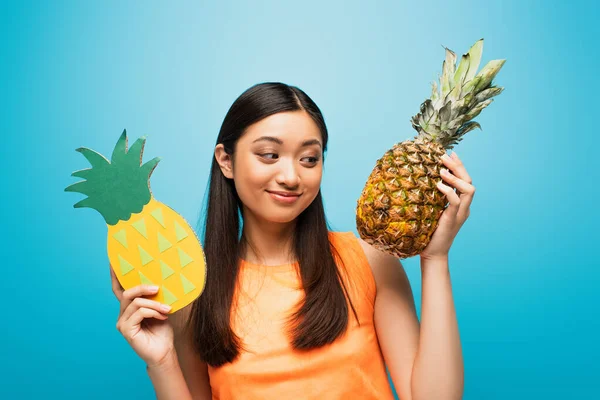 Happy asian girl holding ripe and carton pineapples on blue — Stock Photo