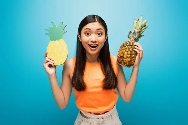 Возбужденная азиатская девушка держит спелые и картонные ананасы на голубом — стоковое фото