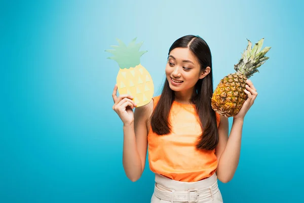 Happy asian girl looking at carton and holding pineapples on blue — Stock Photo