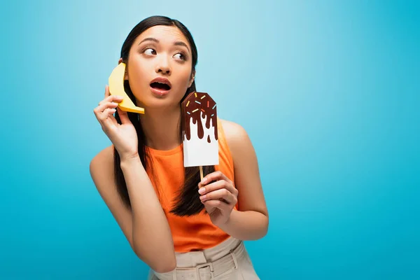 Emotional asian girl with open mouth holding banana near ear and paper ice scream on blue — Stock Photo