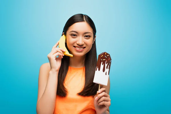 Feliz asiático chica holding banana cerca oreja y papel hielo grito en azul - foto de stock