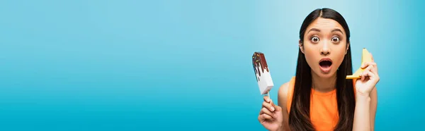 Panoramic crop of shocked asian girl with open mouth holding banana and paper ice scream on blue — Stock Photo