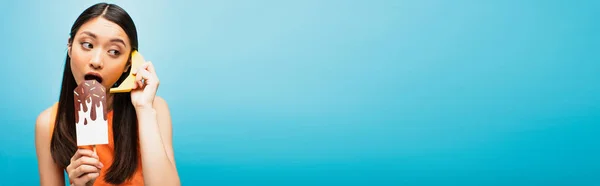 Concetto panoramico di bella ragazza asiatica con banana e ghiaccio di carta urlare sul blu — Foto stock