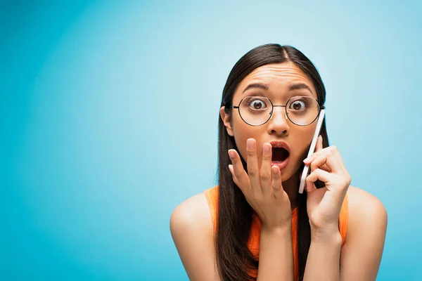 Шокированная азиатская девушка в очках, прикрывающих рот, разговаривая по смартфону в синем — стоковое фото
