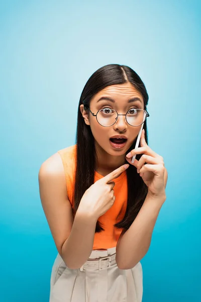 Shocked asian girl in glasses pointing with finger at smartphone while talking on blue — Stock Photo