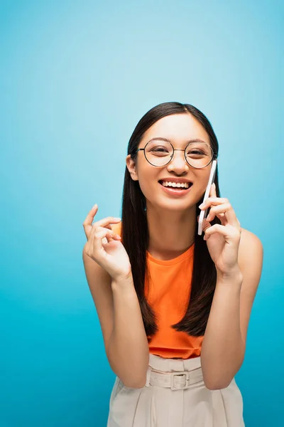 Smiling asian girl in glasses talking on smartphone on blue — Stock Photo