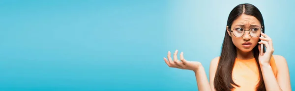 Panoramic crop of displeased asian girl in glasses talking on smartphone and gesturing on blue — Stock Photo