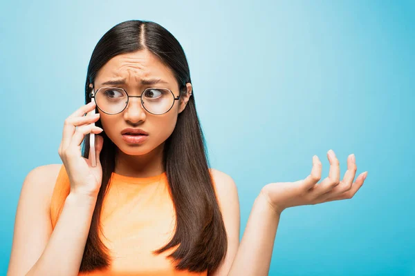 Незадоволена азіатська дівчина в окулярах говорить на смартфоні і жестикулює на синьому — стокове фото