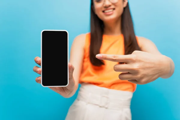 Обрізаний вид щасливої дівчини, що вказує пальцем на смартфон з порожнім екраном ізольовано на синьому — стокове фото
