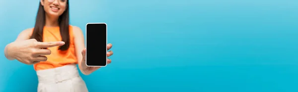 Panoramic crop of happy girl pointing with finger at smartphone with blank screen isolated on blue — Stock Photo