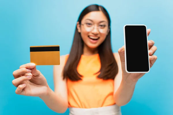 Enfoque selectivo de la chica asiática feliz celebración de teléfono inteligente con pantalla en blanco y tarjeta de crédito aislado en azul - foto de stock