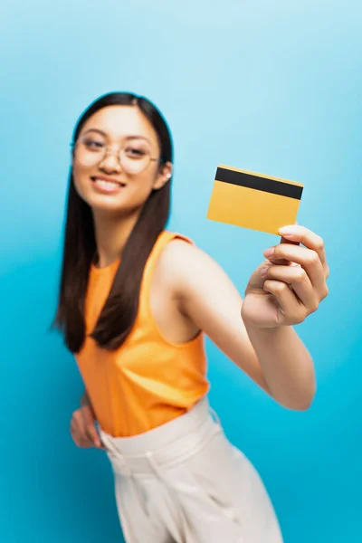 Enfoque selectivo de feliz chica asiática en gafas celebración de la tarjeta de crédito en azul - foto de stock