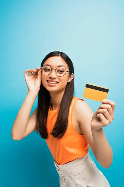 Enfoque selectivo de alegre chica asiática en gafas celebración de la tarjeta de crédito en azul - foto de stock