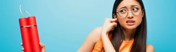 Panoramic crop of stressed asian girl in glasses covering ear and holding dynamite sticks on blue — Stock Photo